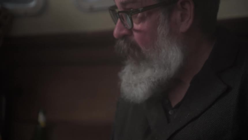A man with a long beard and glasses is looking down as he sits on a couch