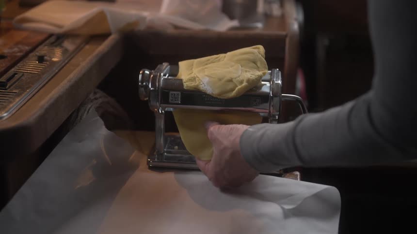 Older woman rolls pasta dough through a hand cranked pasta machine