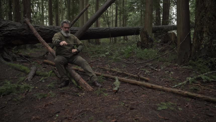Bearded man sitting in the forest making a bow with wooden branches
