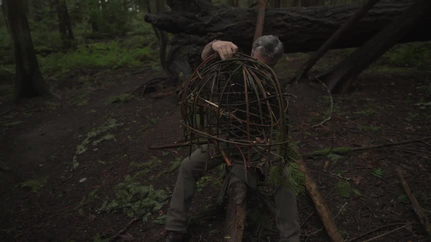 Older man sits on a fallen branch in the woods and examines a cage constructed of thin branches
