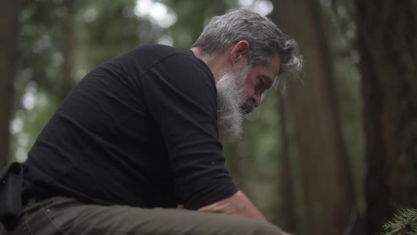 Older man uses a stick while trying to start a fire in the woods during the daytime