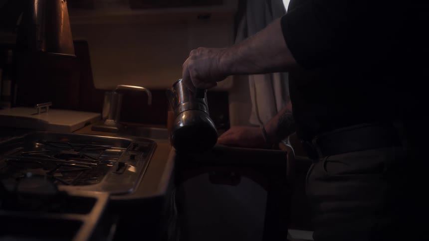 A close up shows a man pouring coffee from an espresso maker into a cup in a dimly lit kitchen