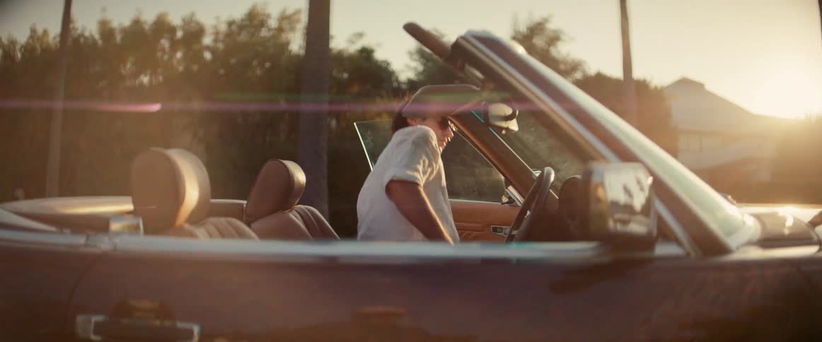 Man wearing sunglasses gets into a convertible with its top down as the sun glares from the horizon