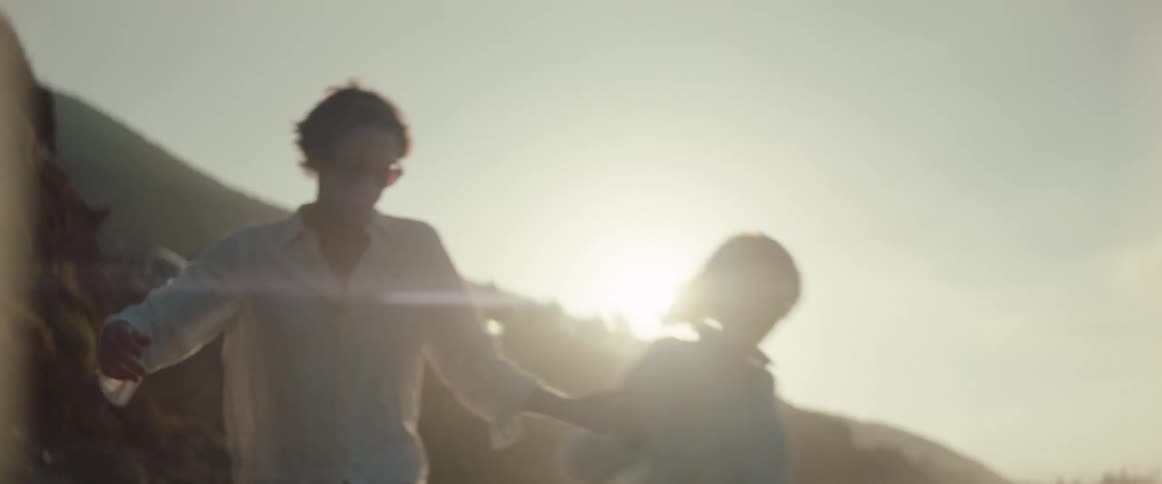 A backlit couple holds hands while walking towards the camera along a sandy beach shoreline near the ocean waves during a bright sunset