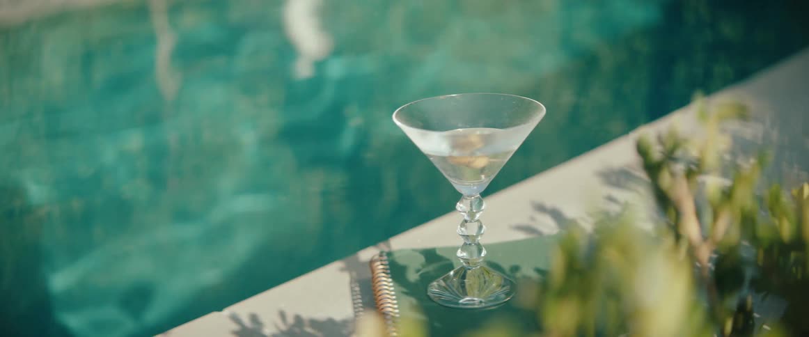 A martini sits on top of a green notebook beside a swimming pool on a sunny day