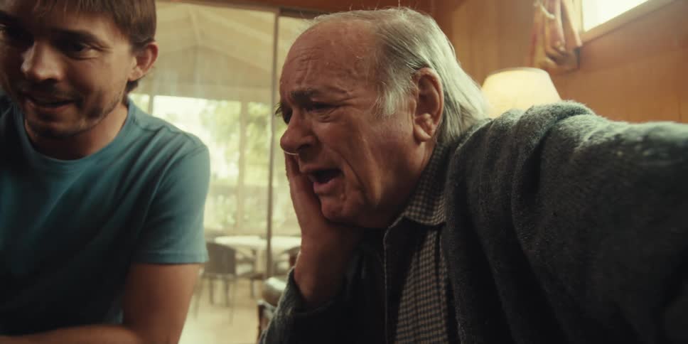 An elderly man sits in his living room while looking at a laptop screen with a younger man