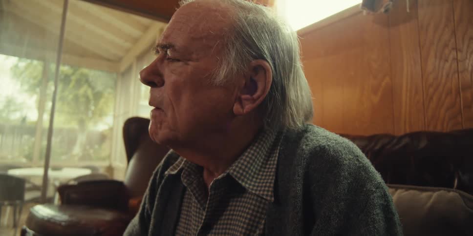 A young man sits beside an elderly man in a cozy home on a sunny day as the elderly man appears puzzled