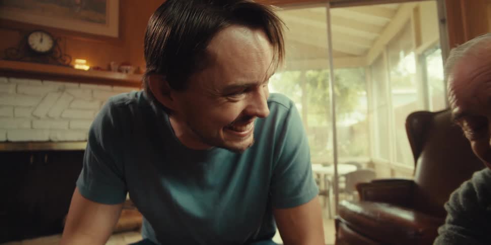 A young man and an elderly man sit together in a cozy home on a sunny day happily laughing while focused on a laptop