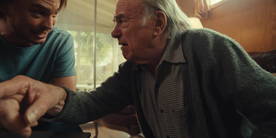 A young man and an elderly man sit together in a cozy home on a sunny day talking while focused on a laptop