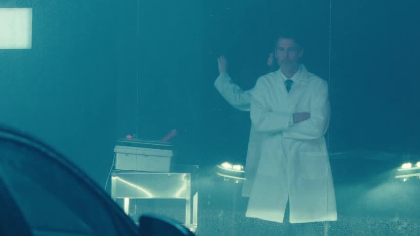 Scientist with arms crossed wearing a white lab coat observing a car with its headlights on