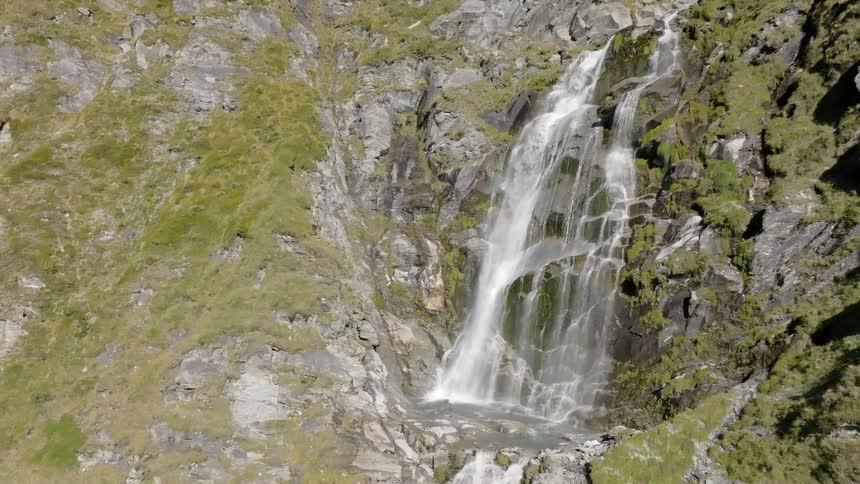 A cascading waterfall flows down a rocky mountainside covered in sparse vegetation as the view slowly zooms in