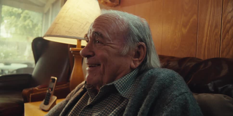 A close up of an elderly man sitting on a couch at home with a placid smile as he talks to someone