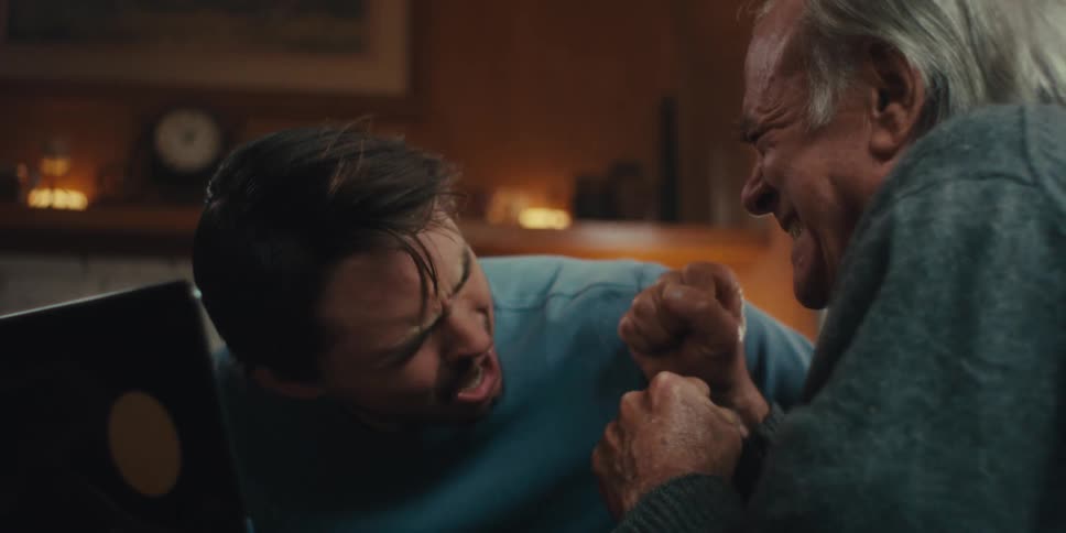 A younger man and an older man watch something intensely on a screen erupting into simultaneous contrasting reactions of excitement and dismay