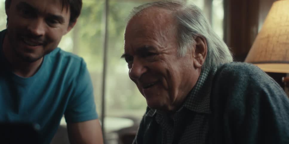 A younger man and an older man share a moment of joy and amusement looking at a laptop initially waving before the focus shifts closely to each of their smiling faces