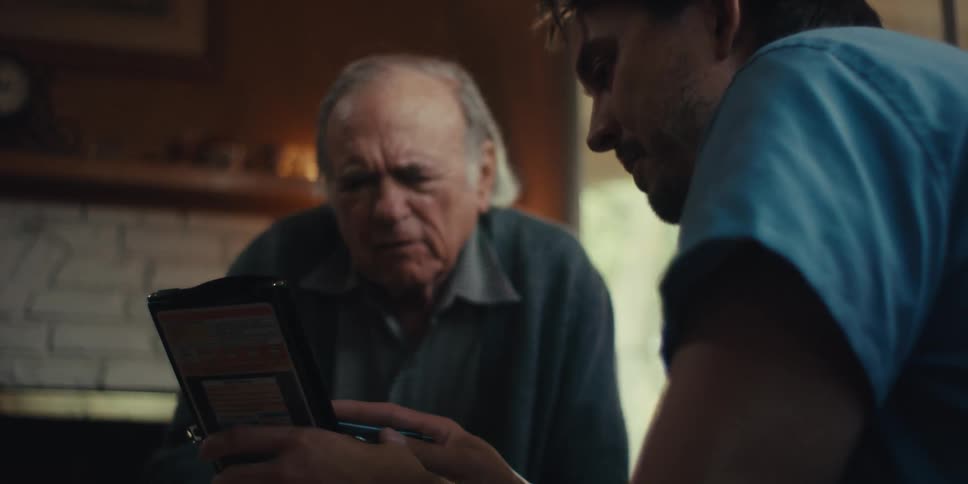 In a dimly lit room an older man intently studies a handheld device held by a younger man beside him