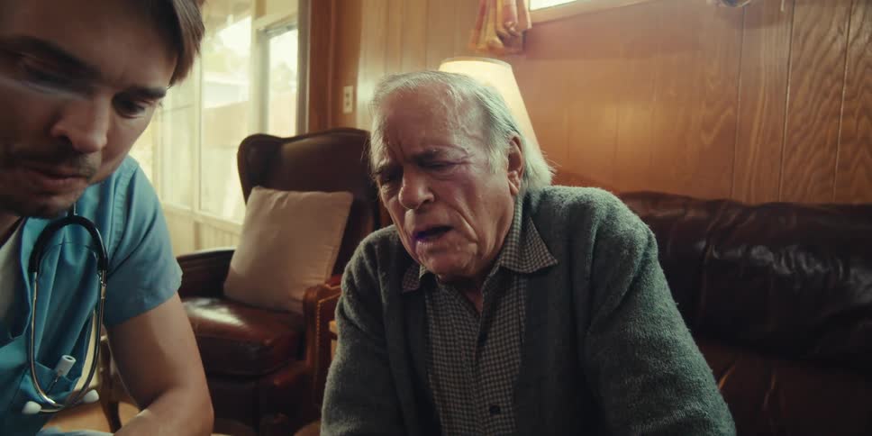A nurse reads a medical chart next to an elderly man seated on a couch at home