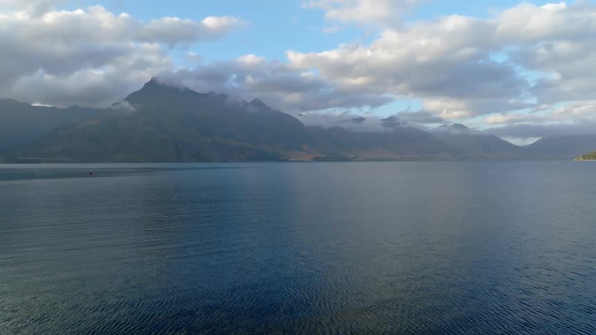 A wide expanse of dark water stretches towards a backdrop of hazy rugged mountains under a sky with scattered clouds