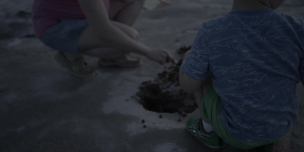 A boy and woman crouch and play in dirt at dusk
