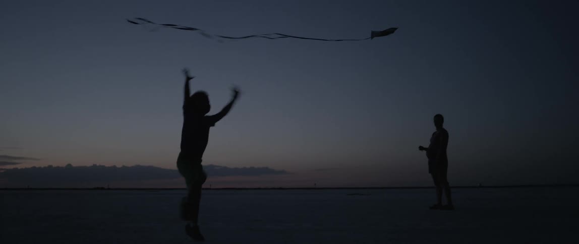 A young boy plays as his father flies a kite on a beach at dusk