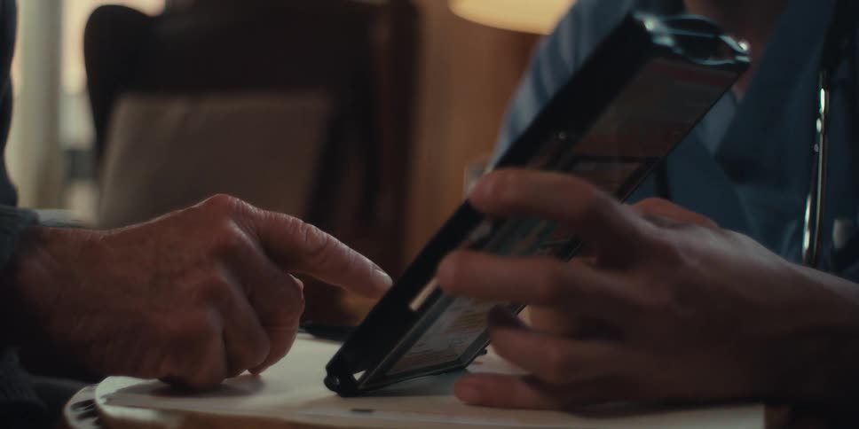 A close up of an elderly man pointing at a medical chart held by a nurse in a home setting