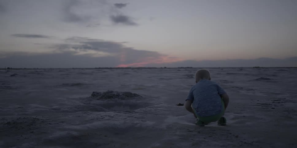 A young boy crouches in the middle of a dirt field at dusk