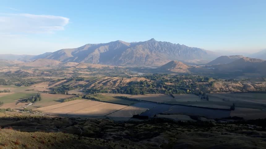 Extensive plateau with agricultural fields and hills in the background dominated by an imposing mountain range under a clear sky