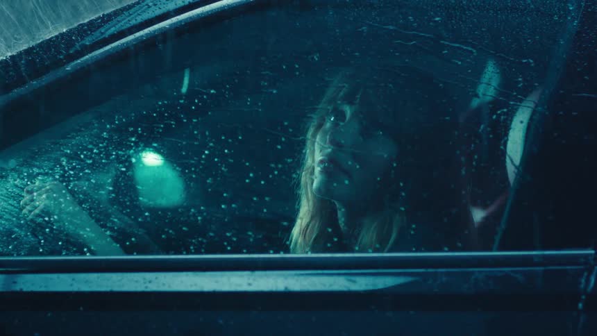 A woman sits in her car and looks out the window during a nighttime rainstorm