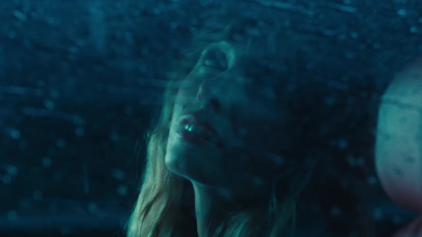 A womans face illuminated by cool blue light looks upward through a rain streaked window at night with bokeh highlights visible