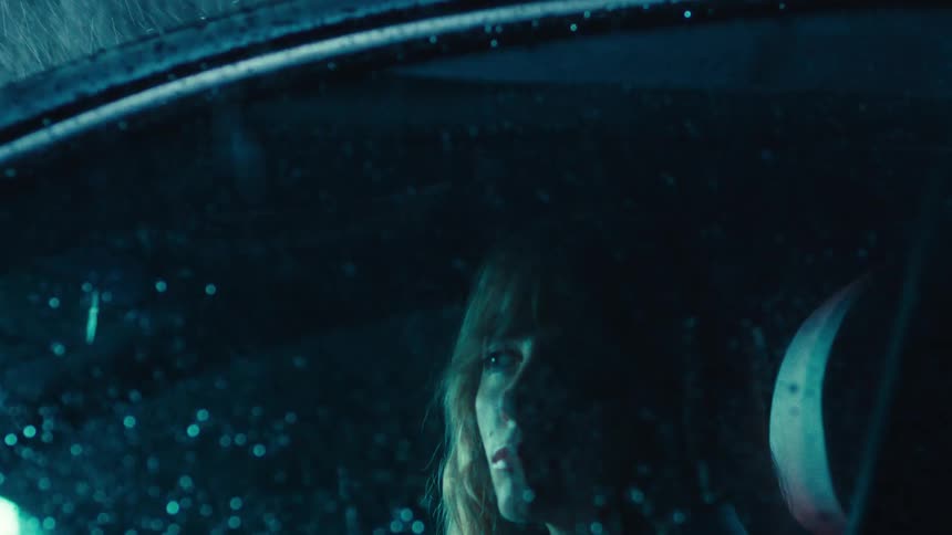 A closeup of a woman sitting in the drivers seat gazing through a rolledup window as heavy rain pours outside at night