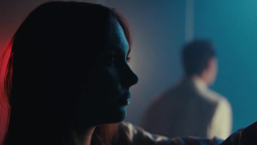 A closeup of a woman in a lab coat turning toward flashing lights illuminating a window while a man in a lab coat gazes out in a dimly lit room