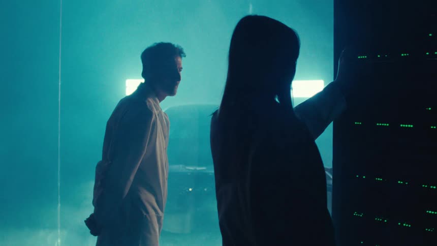 Two scientists in lab coats stand in a room with greenlit lab equipment while strobing lights and smoke surround a vehicle in an adjacent room separated by glass panes