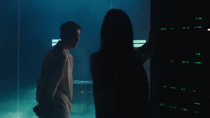 Two scientists in lab coats stand in a room with greenlit lab equipment while strobing lights and smoke surround a vehicle in an adjacent room separated by glass panes