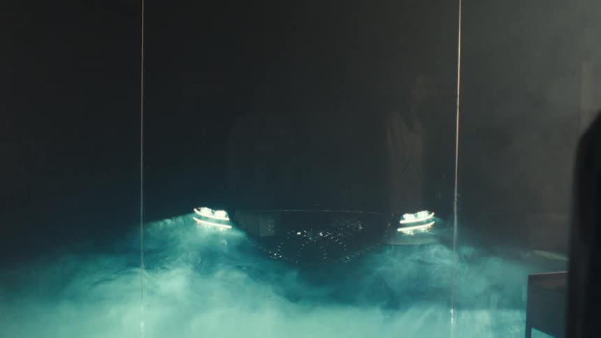 Two scientists in lab coats stand in a room with greenlit lab equipment while lights and smoke surround a vehicle in an adjacent room separated by glass panes