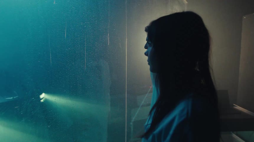Two scientists in lab coats stand in a room while strobing lights and smoke surround a vehicle in an adjacent room separated by glass panes