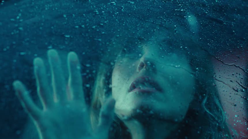 A closeup of a woman sitting in the drivers seat gazing through a rolledup window as heavy rain pours outside at night
