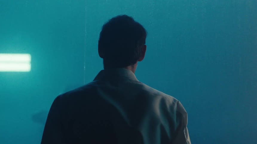A person in a lab coat stands in a smoky dimly lit space with a car seen through glass and smoke illuminated by background lights