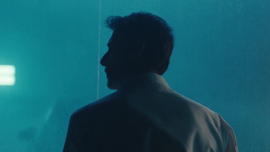 A man in white lab coat stand in a dimly lit room gazing through a misted window at a car enveloped in smoke
