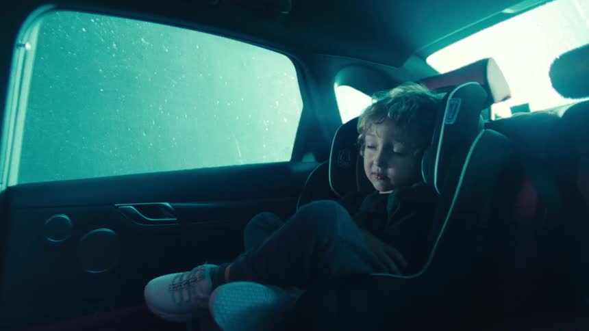 A young child sits in a car seat inside a dimly lit vehicle at night as rain falls and strobing lights illuminate the outside