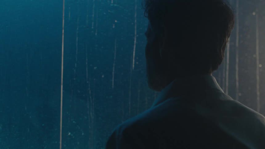 A man in a lab coat is staring out a window on a rainy night at a car with its headlights on