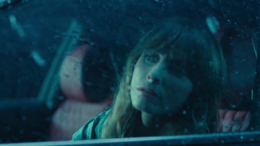 A closeup of a woman sitting in the drivers seat gazing through a rolledup window as heavy rain pours outside at night