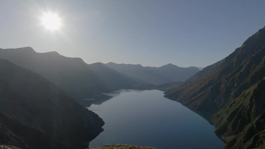 Sunlight illuminates a serene blue lake nestled deep within a valley surrounded by steep shadowy mountains