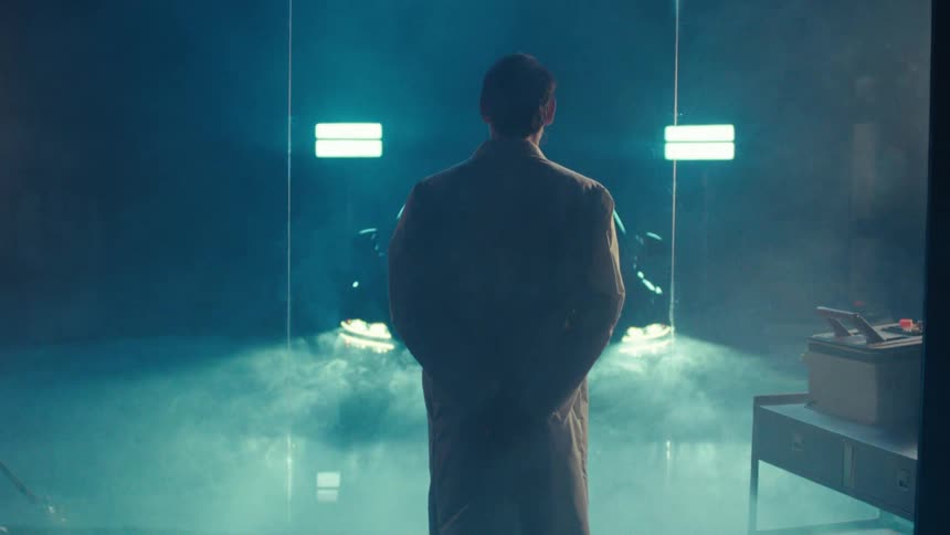 A person in a lab coat stands in a smoky dimly lit space with a cars headlights shining through glass and smoke illuminated by background lights
