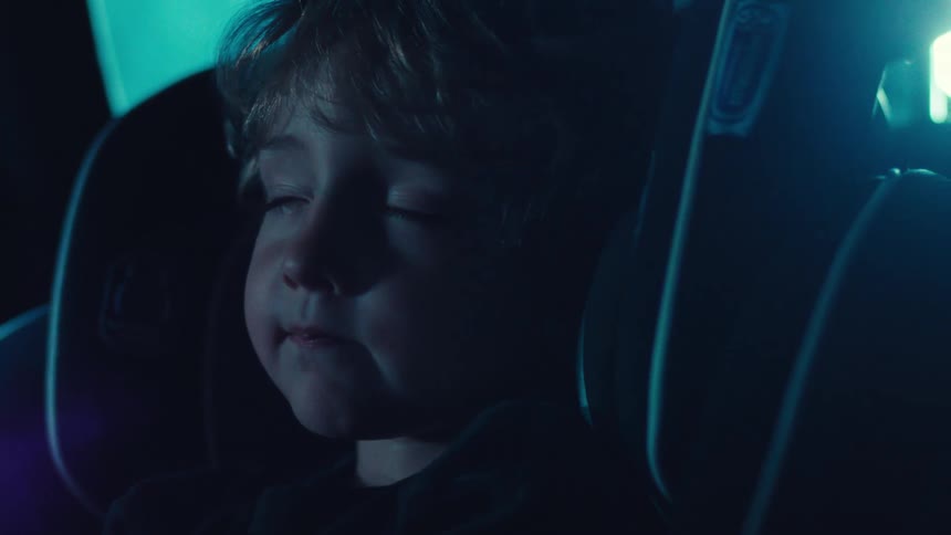 A young child sits in a car seat inside a dimly lit vehicle as rain falls outside at night