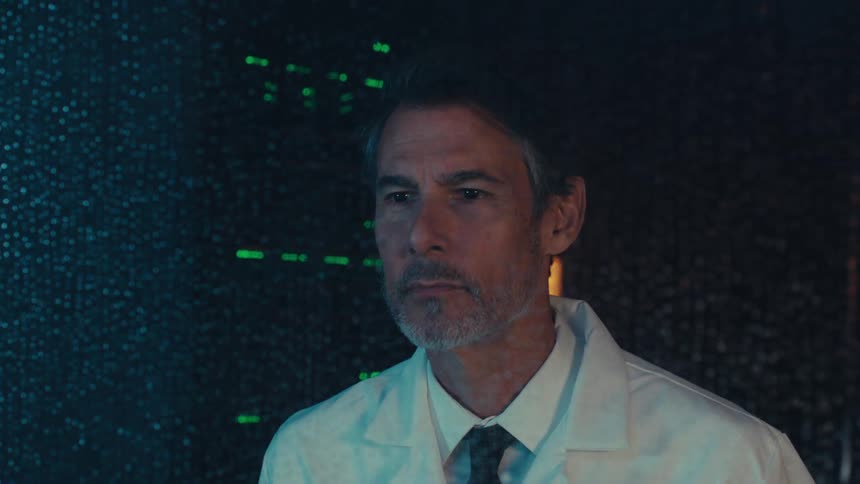 Man in white coat and tie behind glass watching