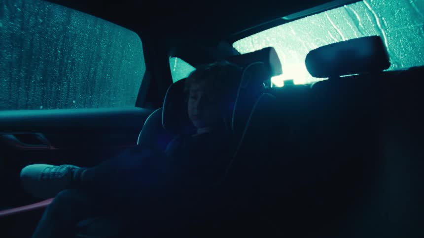 A small child is sleeping in a car seat in the back seat of a car while there is a lightning storm and rain on the vehicle windows