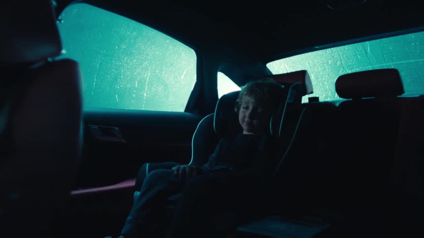 A young child sits in a car seat inside a dimly lit vehicle as rain falls outside at night