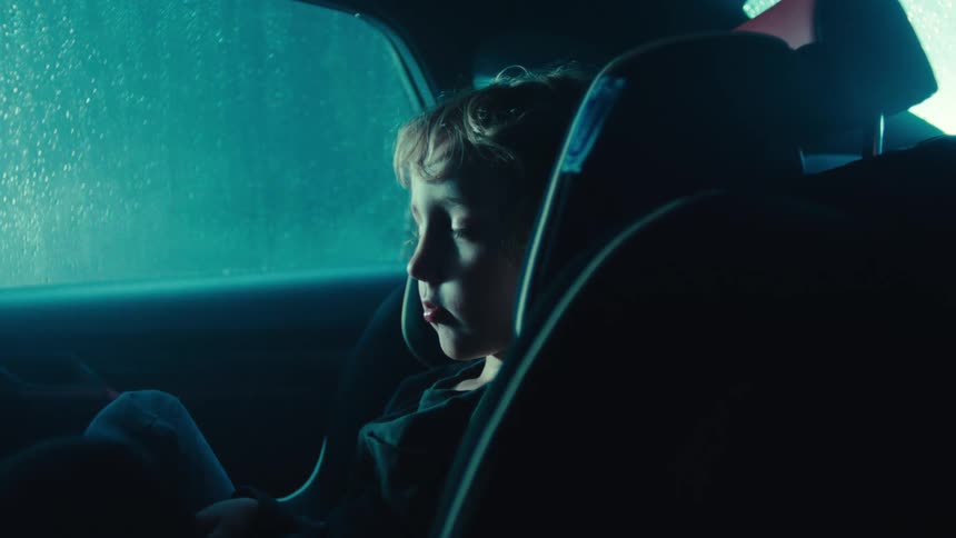 A young child sleepy and talking sits in a car seat inside a dimly lit vehicle as rain falls outside at night