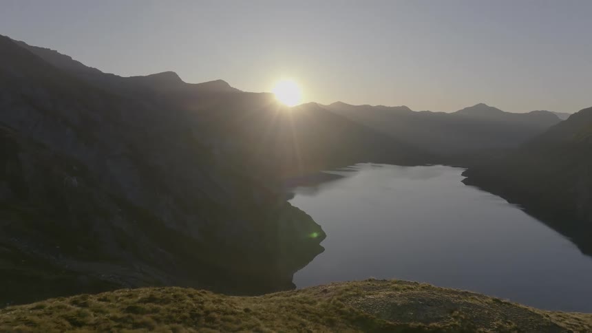 The sun rises over a tranquil lake nestled between steep rocky mountains casting a bright glare on the waters surface