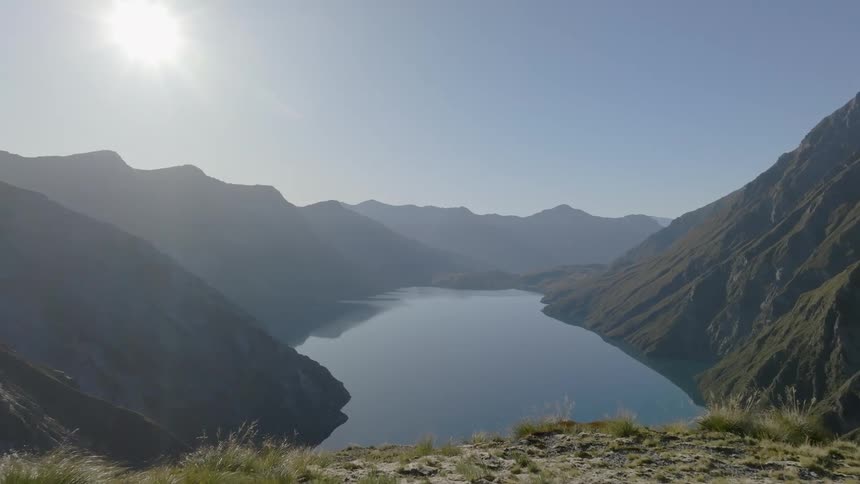 Sunlight illuminates a serene blue lake nestled deep within a valley surrounded by steep shadowy mountains