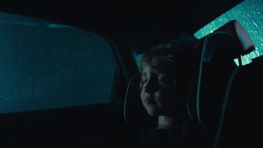 A young child sits in a car seat inside a dimly lit vehicle as rain falls outside at night
